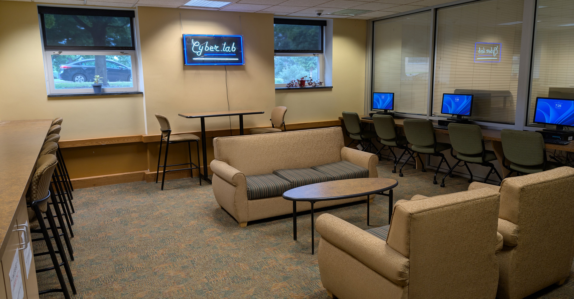 Modern computer lab with collaborative seating area featuring beige upholstered chairs and sofa around a dark coffee table, high-top table with stools near windows, and a row of desktop computers along the right wall. A wall-mounted screen displays 'Cyber lab' and large windows provide natural light.