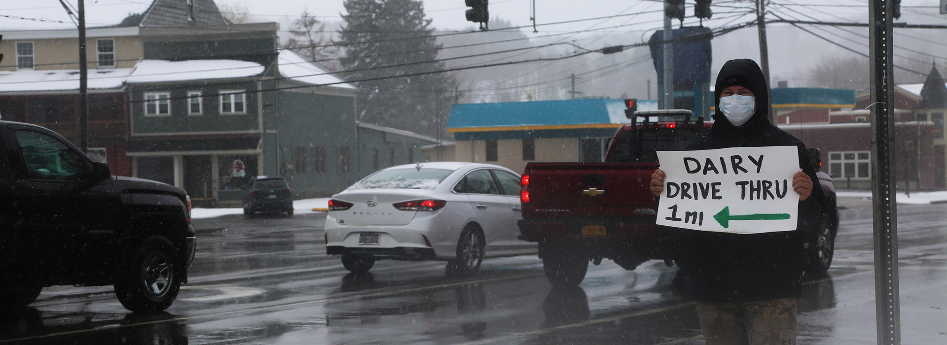 Person wearing a mask on a street corner, holding a sign pointing to a dairy drive