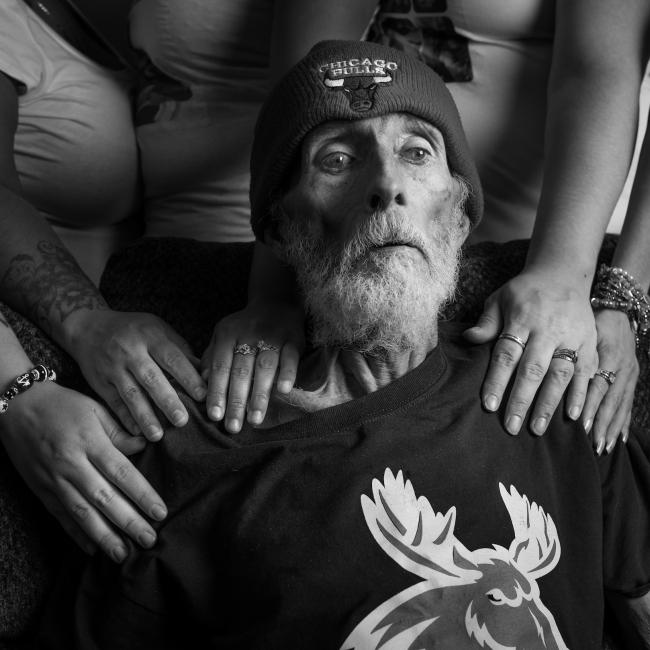 A man with a drawn face and beard with a Chicago Bulls beanie sits with the hands of his daughters on his shoulders