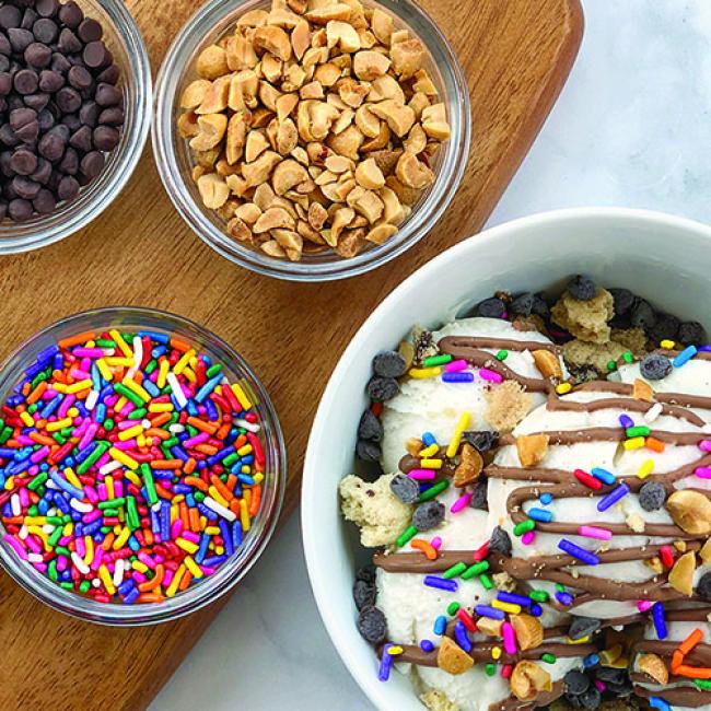 Ice cream sundae bit bowls full of sprinkles, nuts, and chocolate chips