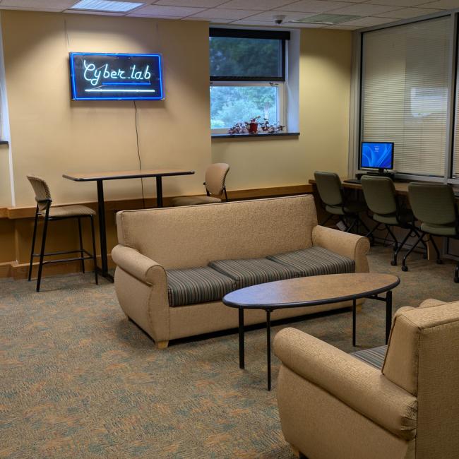 Modern computer lab with collaborative seating area featuring beige upholstered chairs and sofa around a dark coffee table, high-top table with stools near windows, and a row of desktop computers along the right wall. A wall-mounted screen displays 'Cyber lab' and large windows provide natural light.