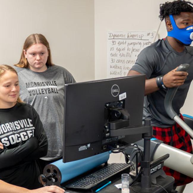 A group of students monitoring a computer while someone exercises on a machine